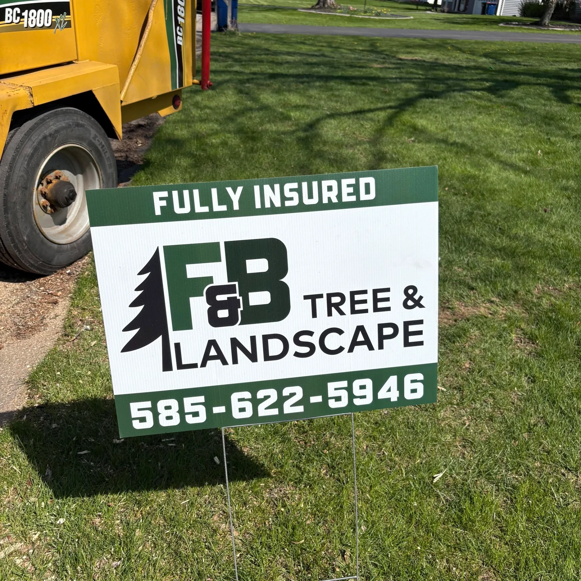 Fully insured F&B Tree and Landscape yard sign staked on a lawn beside the chipper